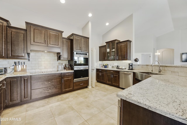 kitchen featuring stainless steel appliances, a sink, dark brown cabinets, light stone countertops, and glass insert cabinets