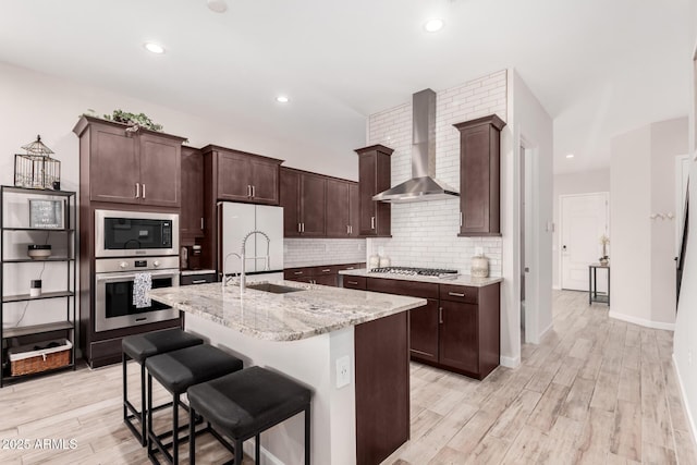 kitchen featuring appliances with stainless steel finishes, wall chimney range hood, light wood-style flooring, decorative backsplash, and a sink