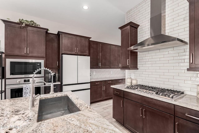 kitchen featuring tasteful backsplash, wall chimney range hood, light stone countertops, appliances with stainless steel finishes, and a sink