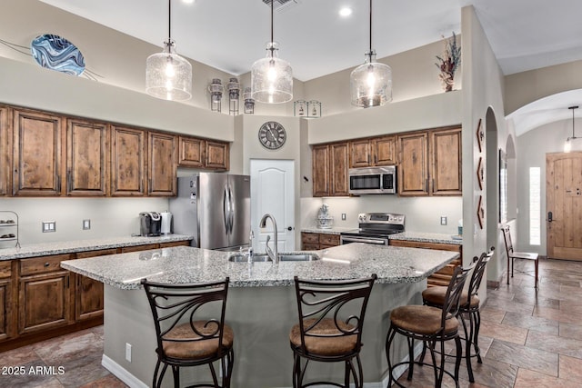 kitchen with arched walkways, stainless steel appliances, a sink, an island with sink, and stone tile flooring