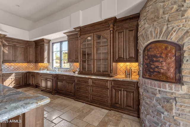 kitchen with stone tile floors, tasteful backsplash, glass insert cabinets, a sink, and light stone countertops