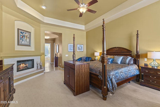 bedroom with baseboards, light carpet, a tray ceiling, a glass covered fireplace, and connected bathroom