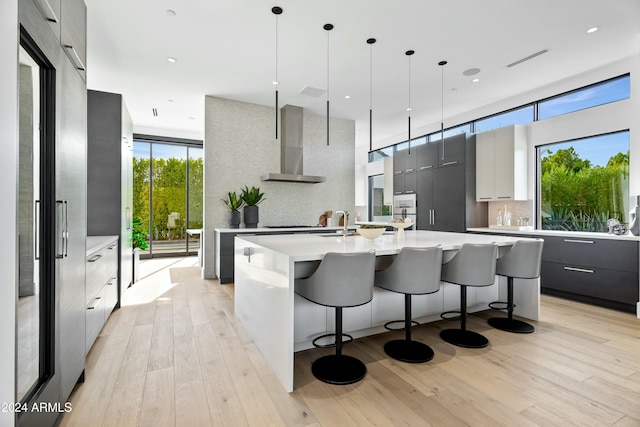 kitchen with an island with sink, pendant lighting, wall chimney range hood, and light hardwood / wood-style floors