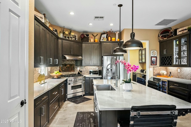 kitchen with light stone countertops, appliances with stainless steel finishes, dark brown cabinetry, decorative light fixtures, and a center island with sink