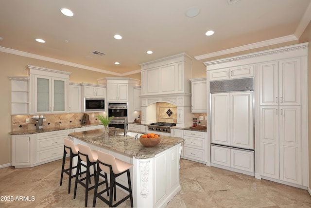 kitchen with built in appliances, an island with sink, white cabinets, and tasteful backsplash