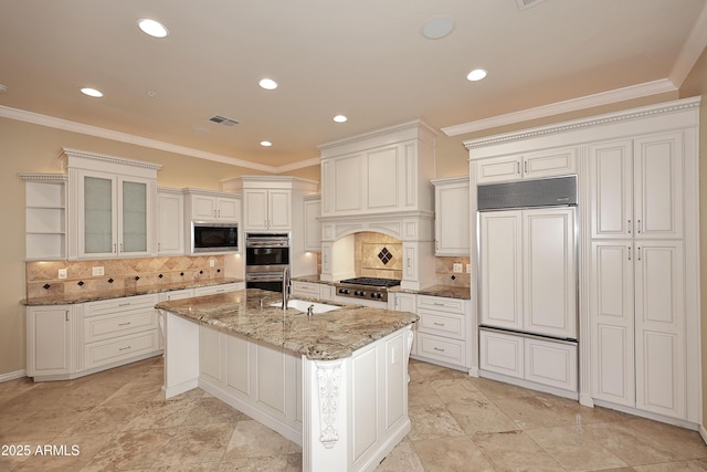 kitchen with built in appliances, white cabinetry, backsplash, light stone counters, and a center island with sink