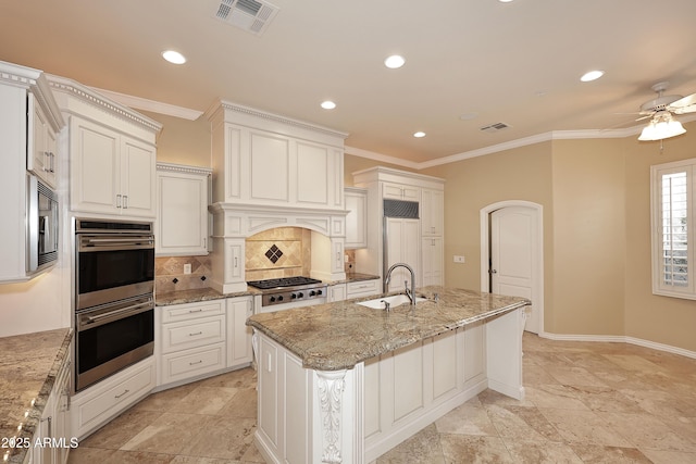 kitchen featuring light stone countertops, built in appliances, an island with sink, sink, and backsplash