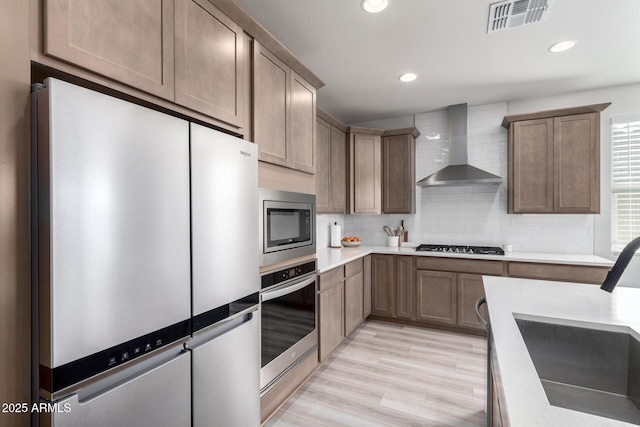 kitchen with wall chimney exhaust hood, light hardwood / wood-style floors, decorative backsplash, appliances with stainless steel finishes, and sink