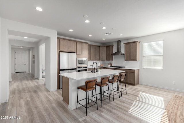 kitchen featuring wall chimney range hood, a kitchen island with sink, light hardwood / wood-style flooring, and appliances with stainless steel finishes