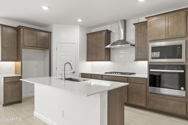kitchen with a kitchen island with sink, backsplash, a sink, stainless steel appliances, and wall chimney exhaust hood