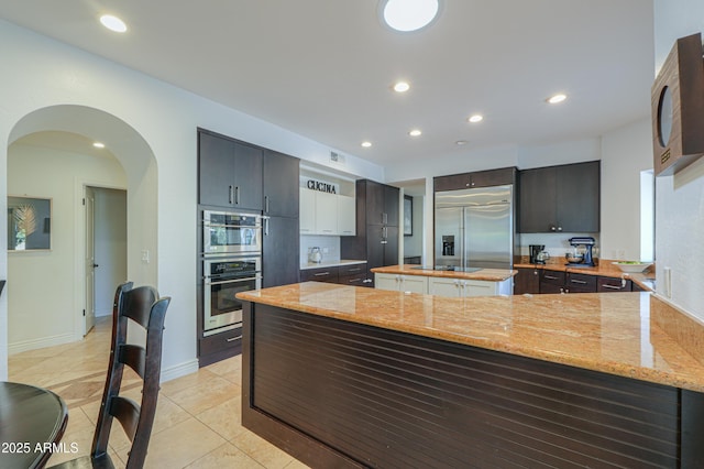 kitchen with kitchen peninsula, stainless steel appliances, light tile patterned flooring, a kitchen island, and light stone counters