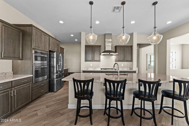 kitchen featuring light wood-type flooring, wall chimney exhaust hood, an island with sink, appliances with stainless steel finishes, and sink