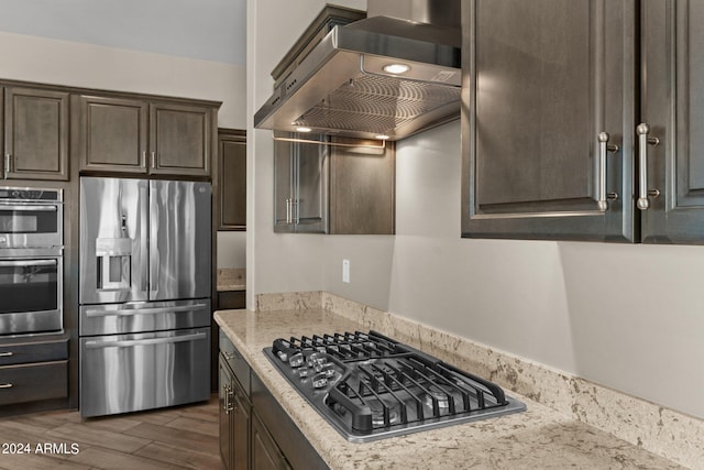 kitchen with wall chimney exhaust hood, dark brown cabinets, light stone countertops, dark hardwood / wood-style floors, and stainless steel appliances