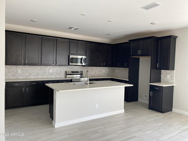 kitchen featuring sink, decorative backsplash, an island with sink, light hardwood / wood-style floors, and stainless steel appliances