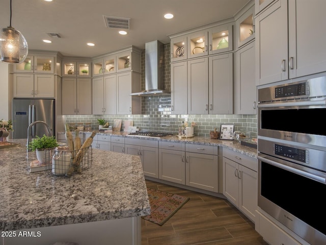 kitchen featuring visible vents, wall chimney exhaust hood, glass insert cabinets, stainless steel appliances, and pendant lighting