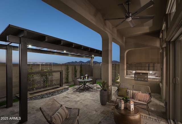 patio terrace at dusk with outdoor dining area, area for grilling, a ceiling fan, a mountain view, and a fenced backyard