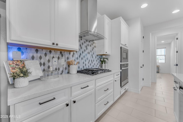 kitchen featuring white cabinetry, stainless steel appliances, wall chimney range hood, light stone counters, and decorative backsplash