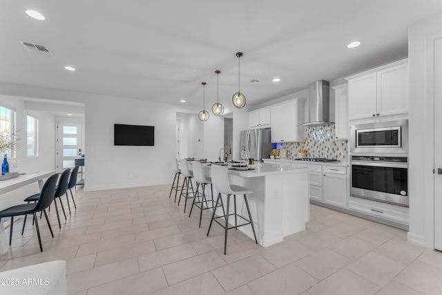 kitchen featuring backsplash, wall chimney exhaust hood, an island with sink, appliances with stainless steel finishes, and white cabinetry