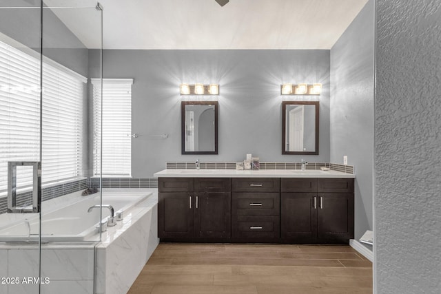 full bathroom with a textured wall, a sink, a bath, wood tiled floor, and double vanity