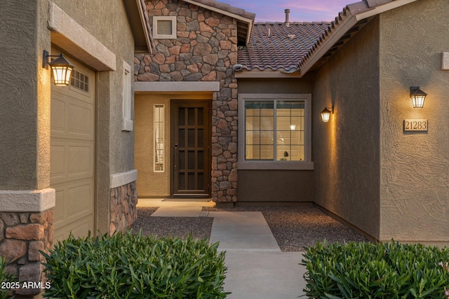 exterior entry at dusk featuring a tiled roof, stone siding, and stucco siding