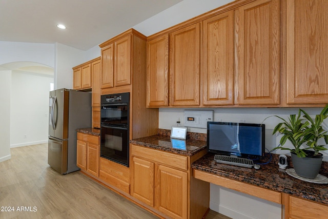 kitchen with dark stone counters, black double oven, light wood-type flooring, built in desk, and stainless steel fridge