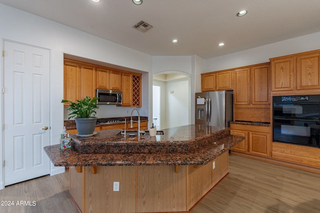 kitchen with dark stone countertops, appliances with stainless steel finishes, a kitchen island with sink, and light hardwood / wood-style flooring