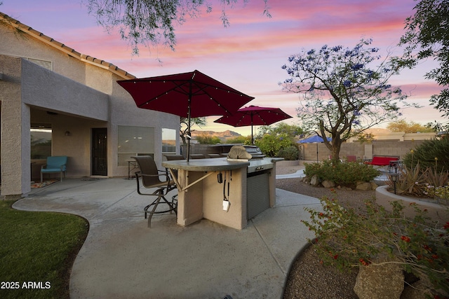 patio terrace at dusk with exterior kitchen, a fenced backyard, outdoor dry bar, and a grill