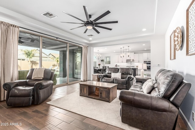 living area featuring dark wood-style floors, ceiling fan with notable chandelier, a raised ceiling, and visible vents