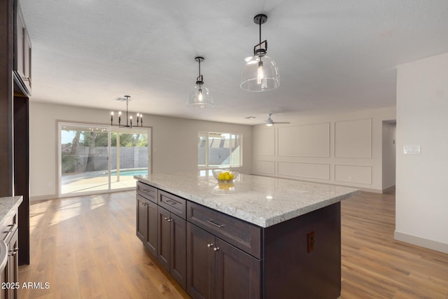 kitchen with dark brown cabinets, hanging light fixtures, a kitchen island, light stone countertops, and light hardwood / wood-style floors