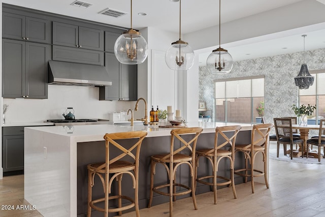 kitchen featuring wall chimney range hood, a breakfast bar, a center island with sink, decorative light fixtures, and light wood-type flooring