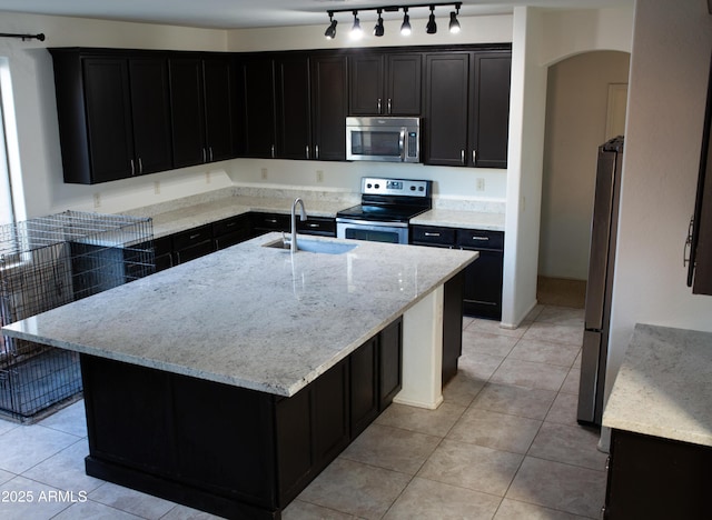 kitchen with sink, light tile patterned floors, appliances with stainless steel finishes, a kitchen island with sink, and light stone counters