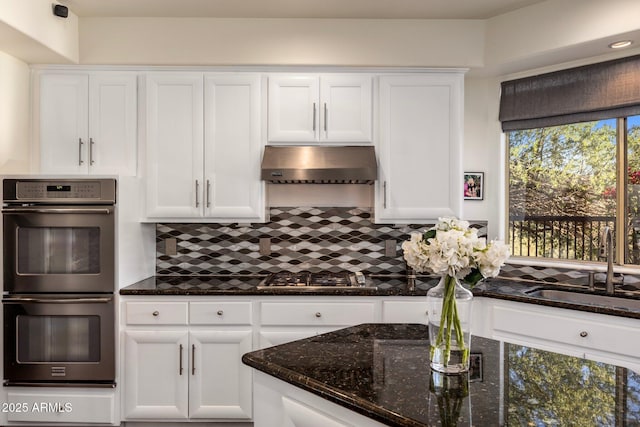 kitchen with dark stone counters, white cabinets, under cabinet range hood, double oven, and stainless steel gas stovetop