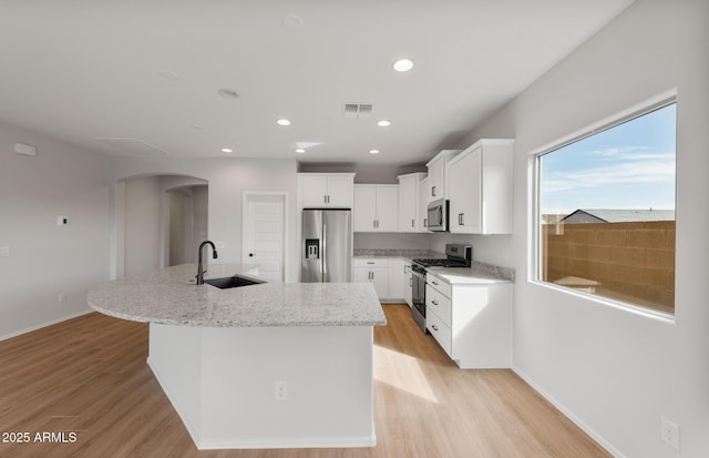 kitchen with arched walkways, a kitchen island with sink, a sink, white cabinetry, and appliances with stainless steel finishes