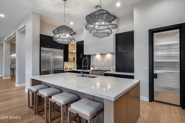 kitchen featuring backsplash, stainless steel built in refrigerator, dark cabinets, light wood-style floors, and a kitchen island with sink