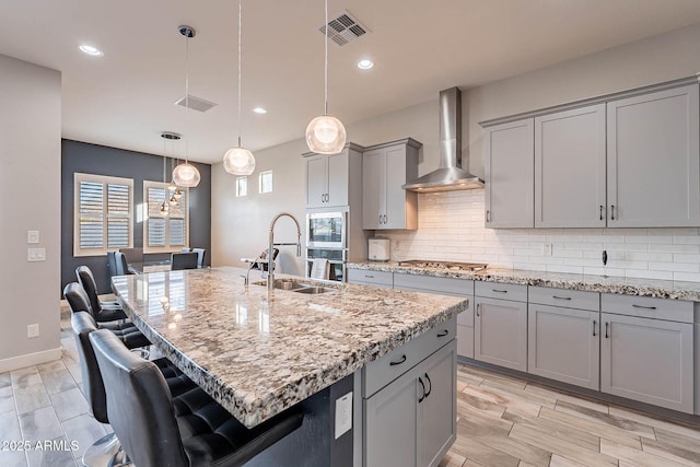 kitchen with a center island with sink, decorative light fixtures, wall chimney exhaust hood, light stone counters, and sink