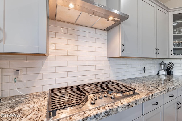 kitchen featuring stainless steel gas stovetop, decorative backsplash, white cabinets, wall chimney exhaust hood, and light stone counters