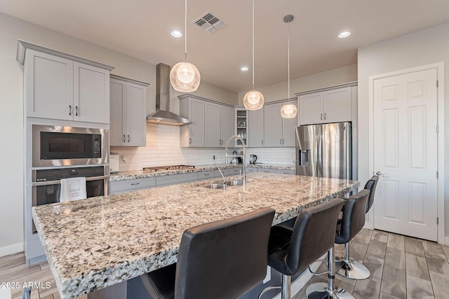 kitchen with wall chimney range hood, an island with sink, sink, and stainless steel appliances