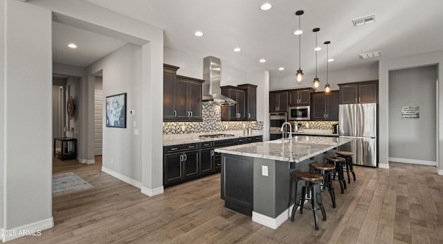 kitchen featuring a kitchen bar, hanging light fixtures, stainless steel appliances, a center island with sink, and wall chimney exhaust hood