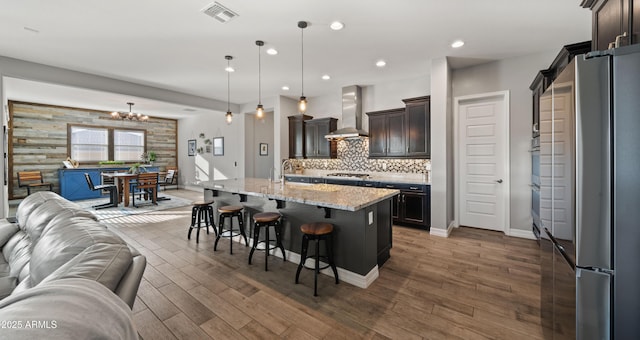 kitchen featuring appliances with stainless steel finishes, a breakfast bar, decorative light fixtures, an island with sink, and wall chimney range hood