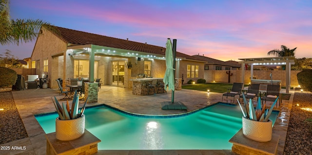 pool at dusk featuring an outdoor kitchen, exterior bar, and a patio