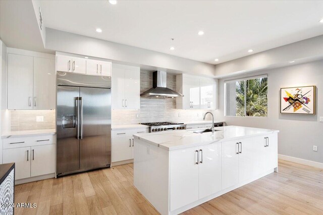 kitchen featuring stainless steel built in refrigerator, white cabinetry, wall chimney exhaust hood, and a kitchen island with sink