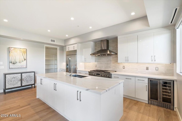 kitchen with a center island with sink, stainless steel appliances, wine cooler, white cabinetry, and wall chimney exhaust hood