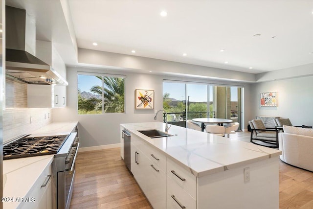 kitchen with sink, a healthy amount of sunlight, a kitchen island with sink, white cabinets, and wall chimney range hood