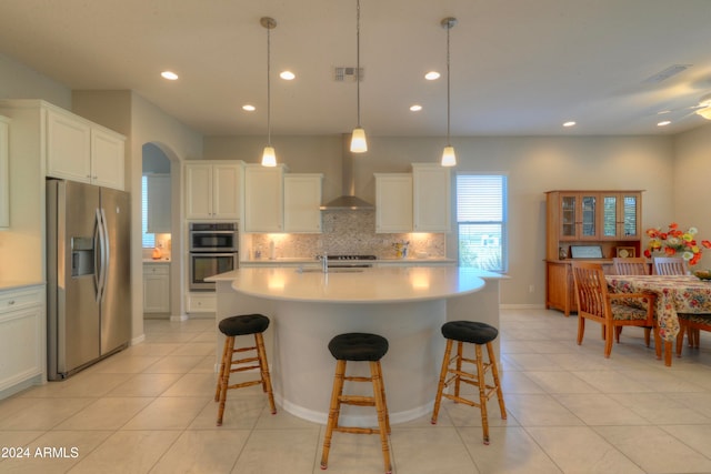 kitchen featuring a center island with sink, hanging light fixtures, appliances with stainless steel finishes, wall chimney range hood, and white cabinets