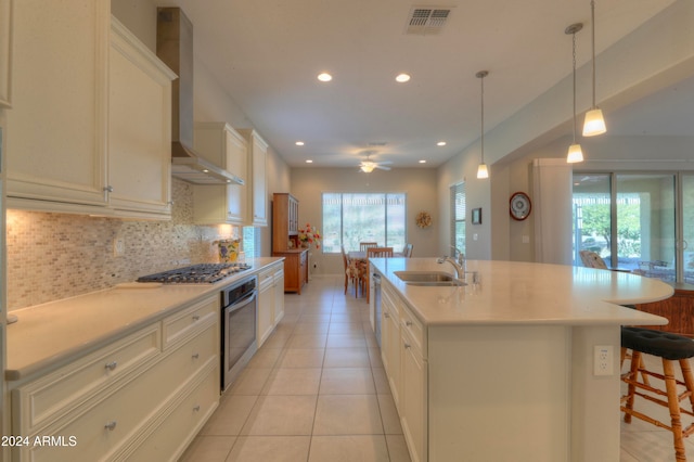 kitchen featuring pendant lighting, decorative backsplash, a kitchen island with sink, stainless steel appliances, and wall chimney range hood