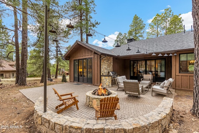 back of house with a patio area, stone siding, board and batten siding, an outdoor fire pit, and a shingled roof
