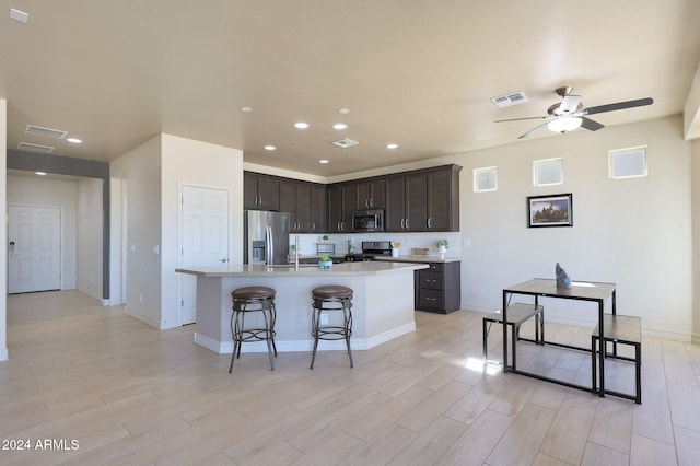 kitchen featuring appliances with stainless steel finishes, light wood-type flooring, dark brown cabinets, ceiling fan, and a kitchen island with sink