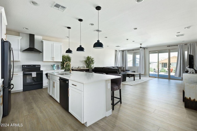 kitchen featuring white cabinetry, wall chimney range hood, a kitchen island with sink, black appliances, and wood-type flooring