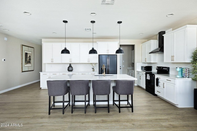 kitchen with wall chimney range hood, an island with sink, light hardwood / wood-style floors, appliances with stainless steel finishes, and a kitchen bar