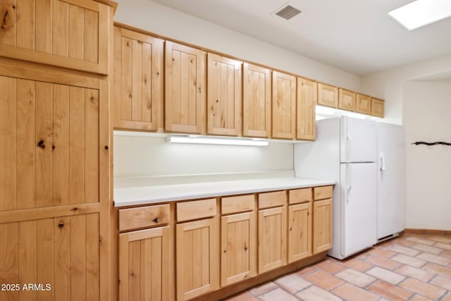 kitchen with visible vents, light brown cabinetry, freestanding refrigerator, light countertops, and brick floor